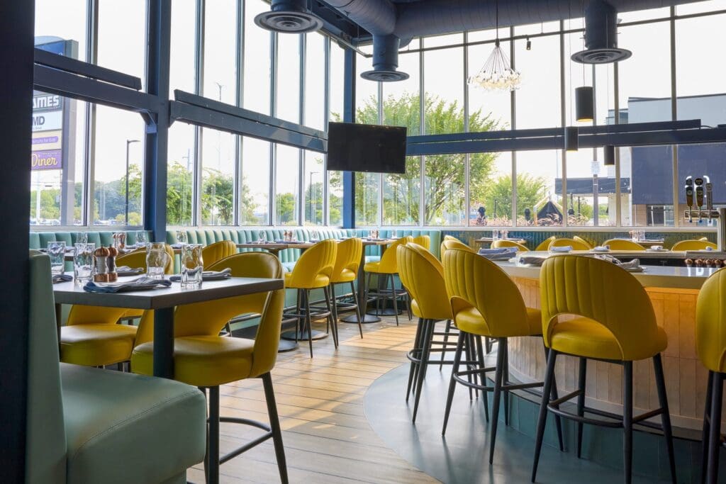 Modern restaurant interior with large windows, yellow chairs around tables and a central bar area. Sunlight streams in, highlighting neatly set tables with glasses and utensils. Outdoor view visible through the glass.