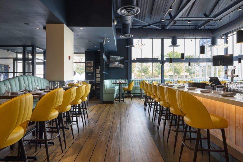 Bright, modern restaurant interior with yellow barstools, wooden floors, and large windows letting in natural light. Tables are set for dining, and contemporary lighting fixtures hang from the ceiling.