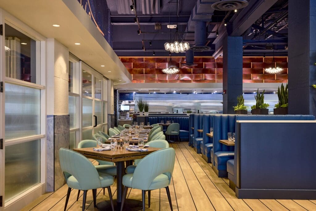 Chic, modern restaurant interior with blue booths, mint green chairs, wooden tables set for dining, light wood floors, plants, and chandeliers under a high ceiling. The kitchen is visible in the background.