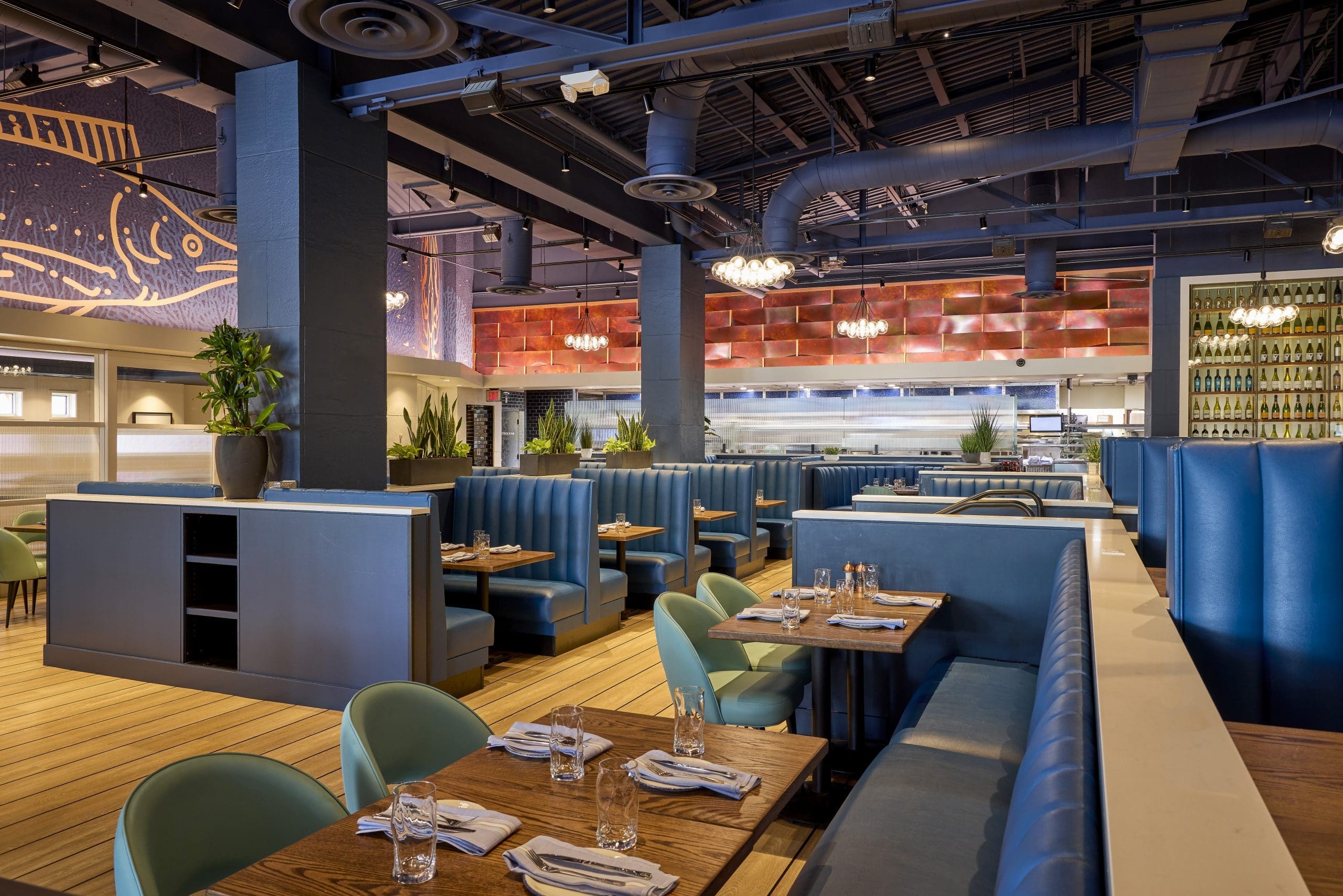 A modern restaurant interior with blue upholstered booths, wooden tables set for dining, green chairs, and exposed industrial ceilings. Decorative plants and art add style, and warm lighting creates a welcoming atmosphere.