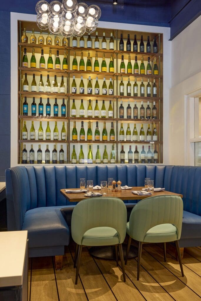 A modern restaurant interior with blue upholstered booth seating, light blue chairs, a wooden table set for four, and a wall display of assorted wine bottles behind glass shelves. A modern chandelier hangs overhead.