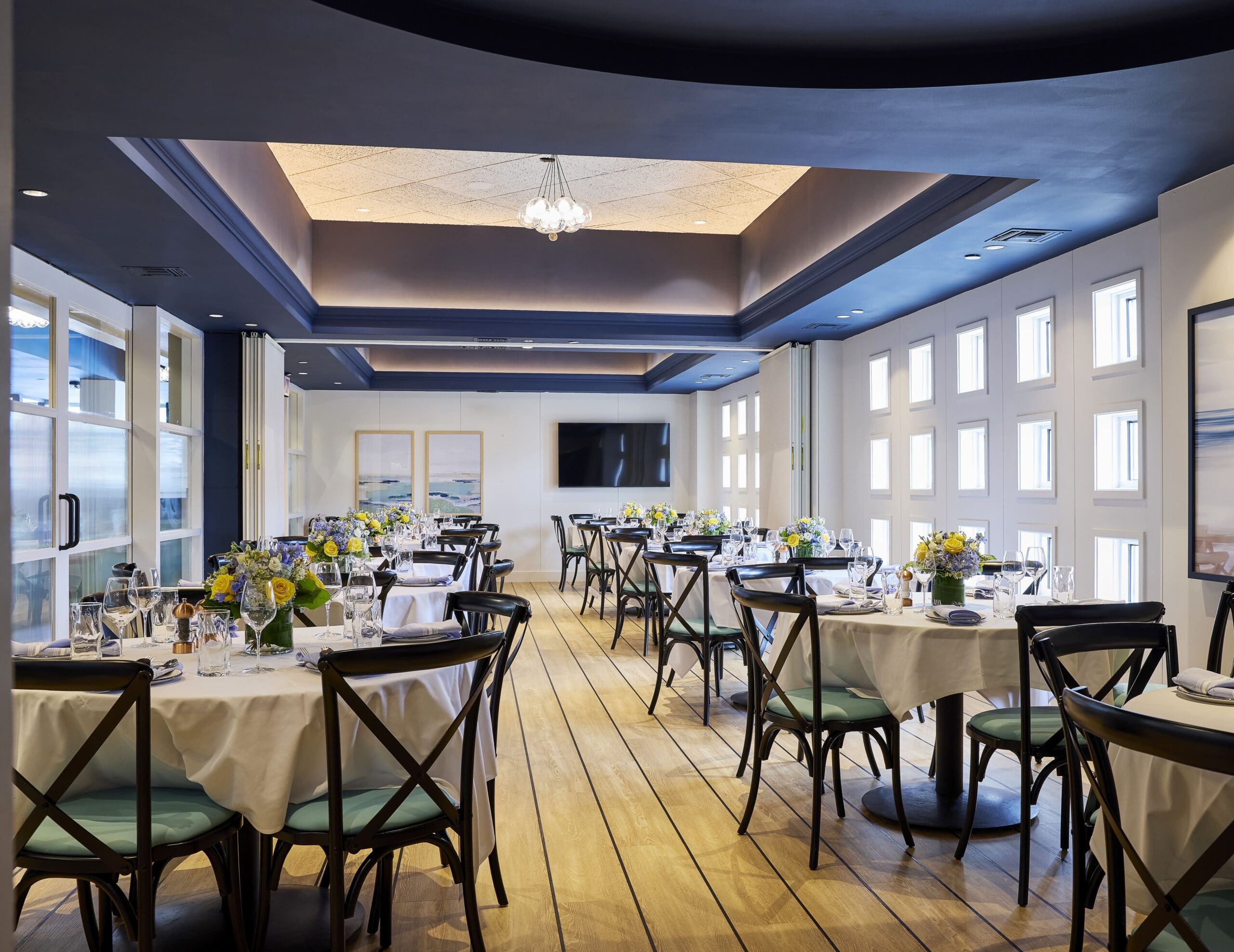 A modern, elegant restaurant dining room with wooden floors, round tables set with white cloths, and black chairs. Bright windows line one wall, and floral centerpieces decorate each table. A TV and framed art are visible in the background.
