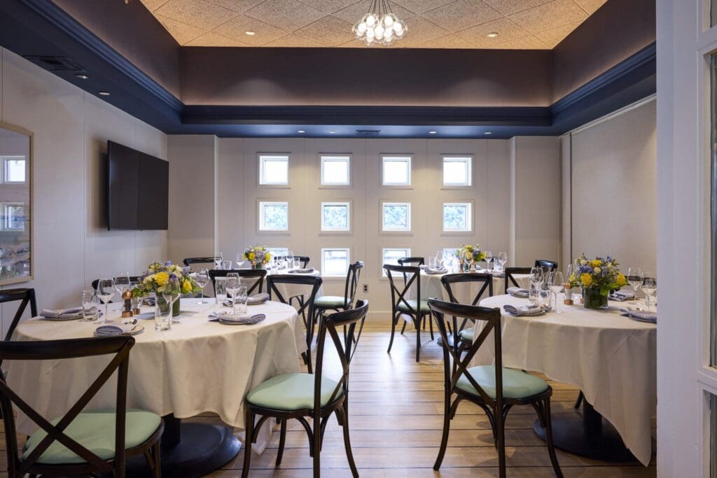 A bright, elegant dining room with round tables set for a meal, decorated with yellow and blue flower centerpieces. Wooden chairs surround the tables, and small square windows line the back wall.