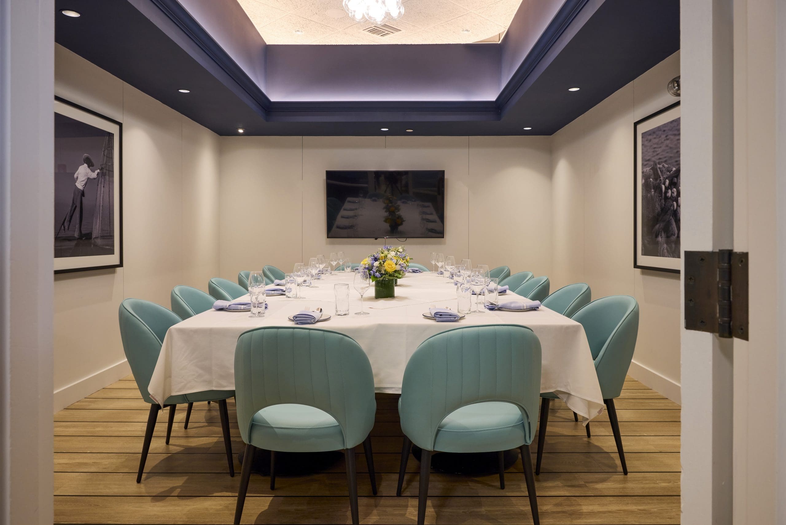 A modern dining room with a long rectangular table set for a meal, surrounded by light blue chairs. White tablecloth, floral centerpiece, and elegant place settings. Artworks hang on white walls; a chandelier hangs from a recessed ceiling.