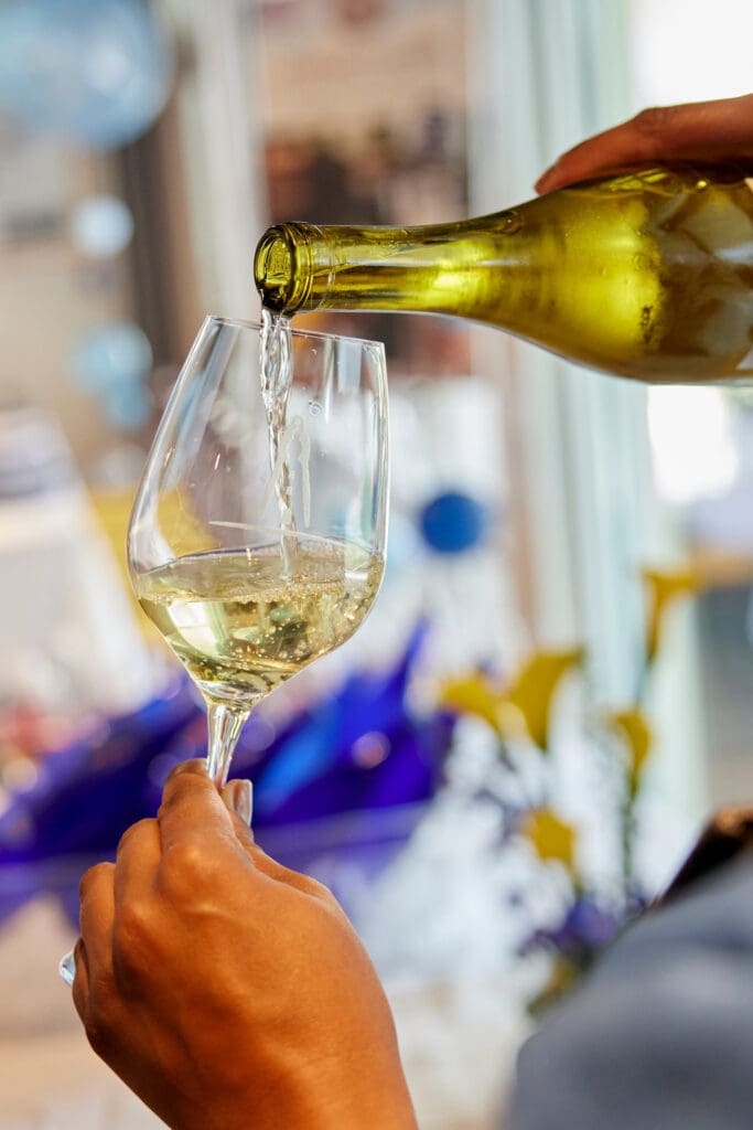 A person pours white wine from a bottle into a wine glass, with a blurred background featuring blue and yellow decorations.