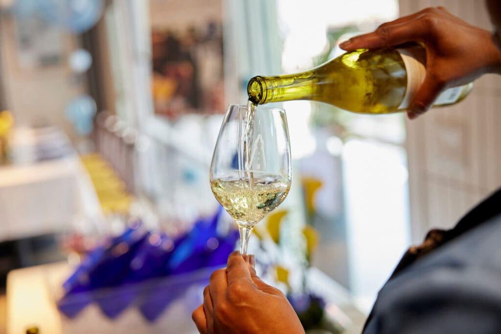 A person pours white wine from a bottle into a clear wine glass at an indoor gathering, with a blurred background of tables and blue decorations.