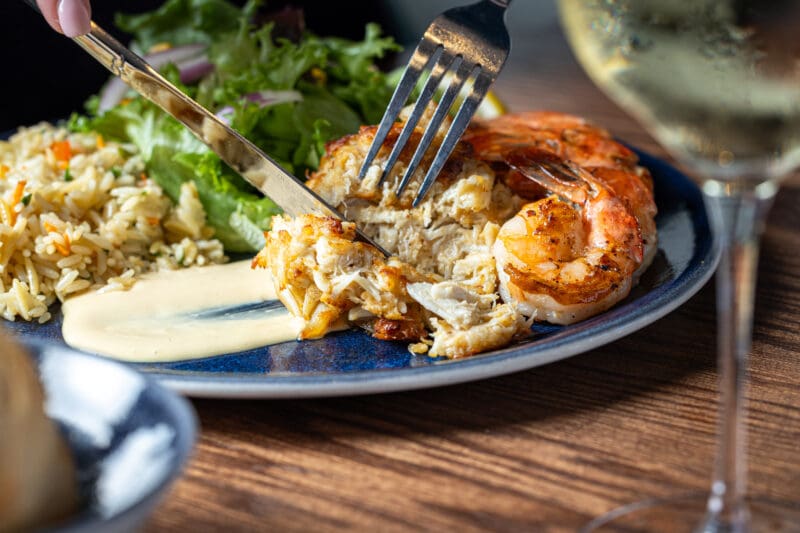 A close-up of a plate with rice, salad, shrimp, and a creamy sauce. A fork and knife are cutting into a piece of crab cake. A glass of white wine is visible on the right side of the image.