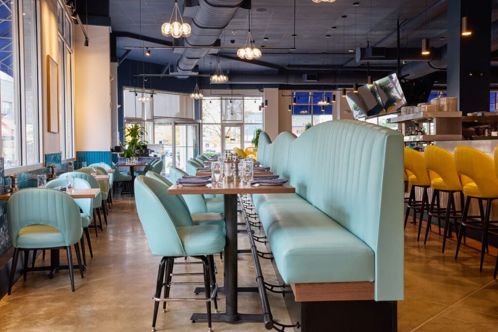 A modern restaurant interior with light blue upholstered booths and chairs, wooden tables set with glasses and napkins, yellow bar stools, large windows, and contemporary lighting. The space is bright and inviting.
