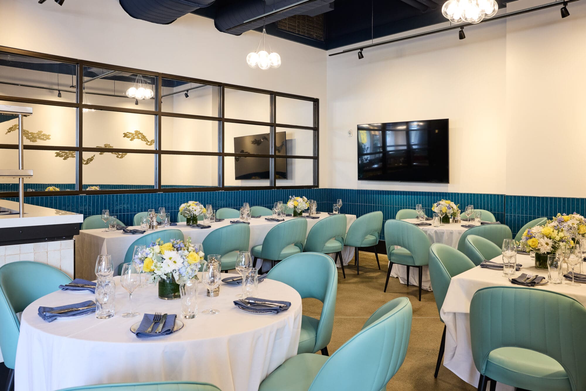 A modern dining room with round tables covered in white cloths, set with blue napkins, glassware, and floral centerpieces. Teal upholstered chairs surround the tables, and a large TV is mounted on a wall with blue paneling.