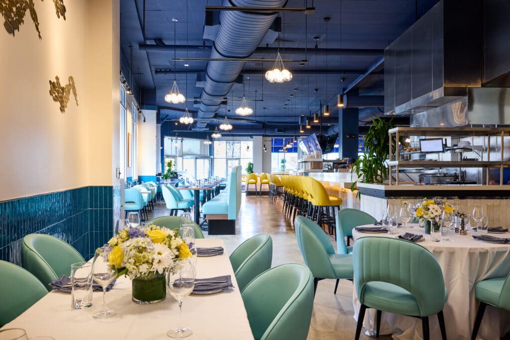 A modern restaurant with teal chairs, white tablecloths, and floral centerpieces. The open kitchen is visible, and a long bar with yellow stools runs along the right side. Large windows provide natural light.