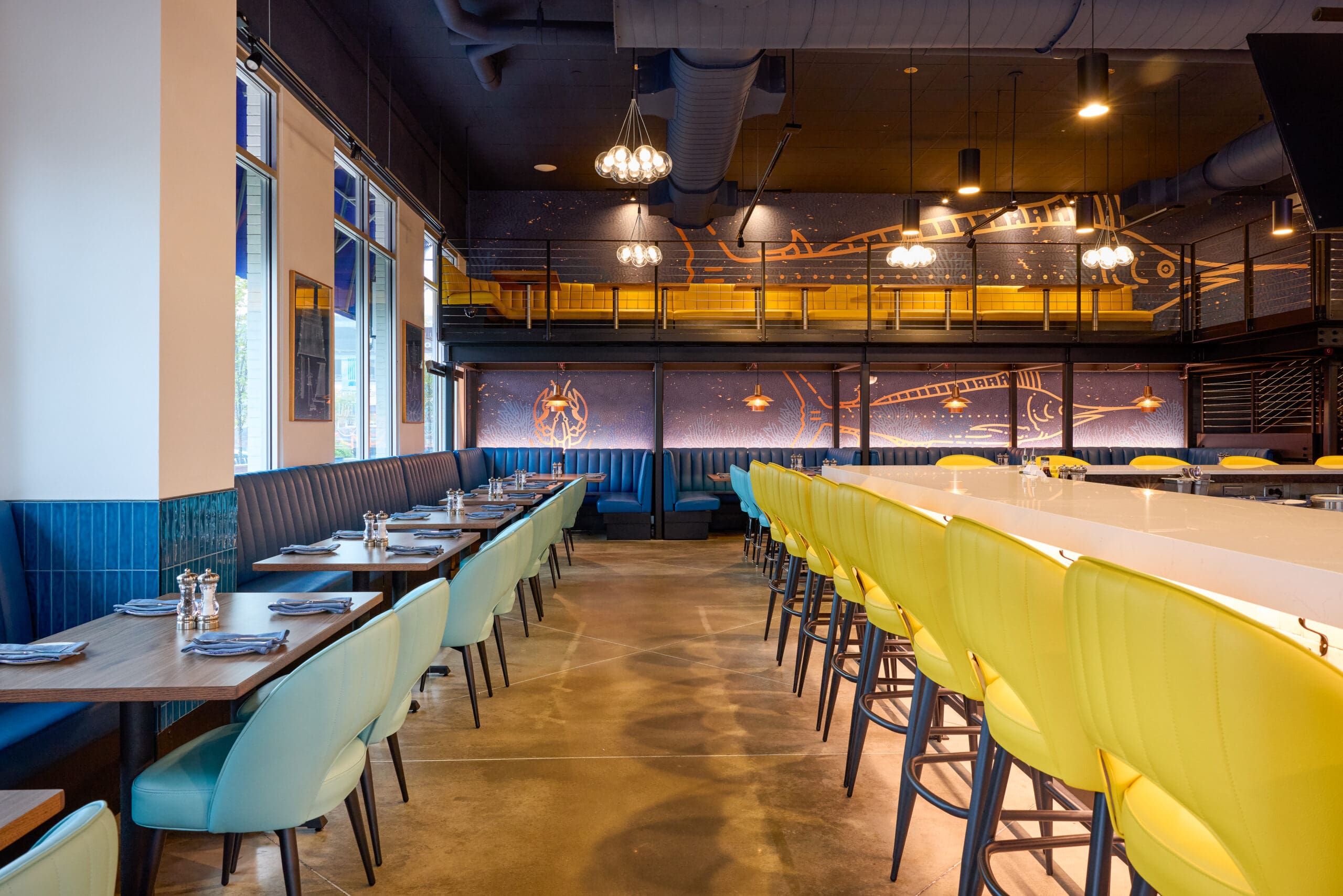 Modern restaurant interior with blue and yellow chairs, tables set with glassware and napkins, long bar with yellow stools, high ceilings, and a mural featuring celestial and aquatic designs on the back wall.