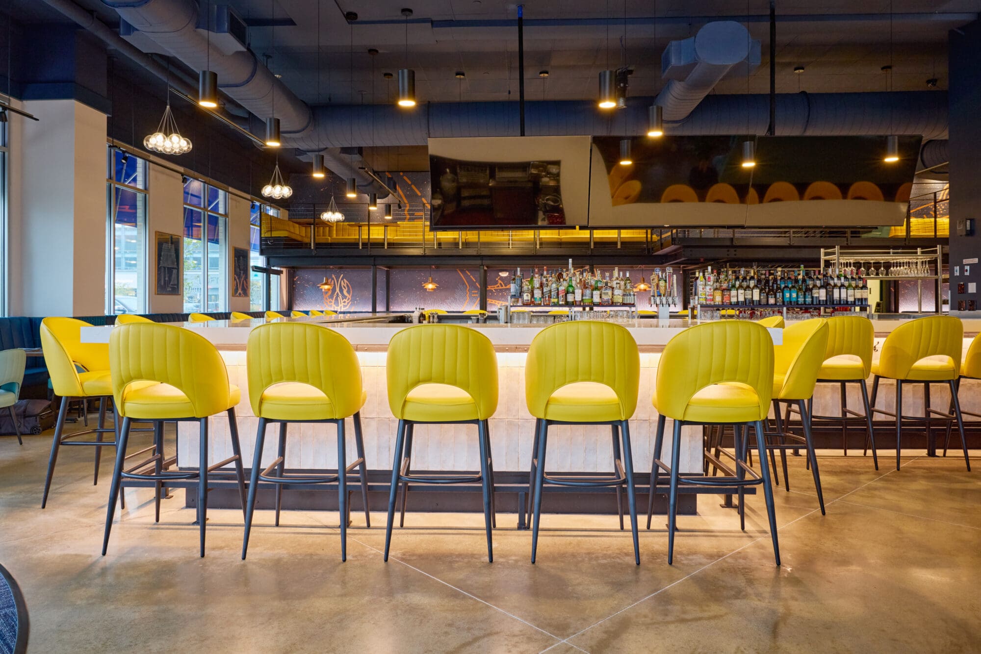 A modern bar with a row of bright yellow chairs lined up at a counter, shelves of liquor bottles behind the bar, pendant lights hanging from the ceiling, and industrial-style decor with exposed pipes.