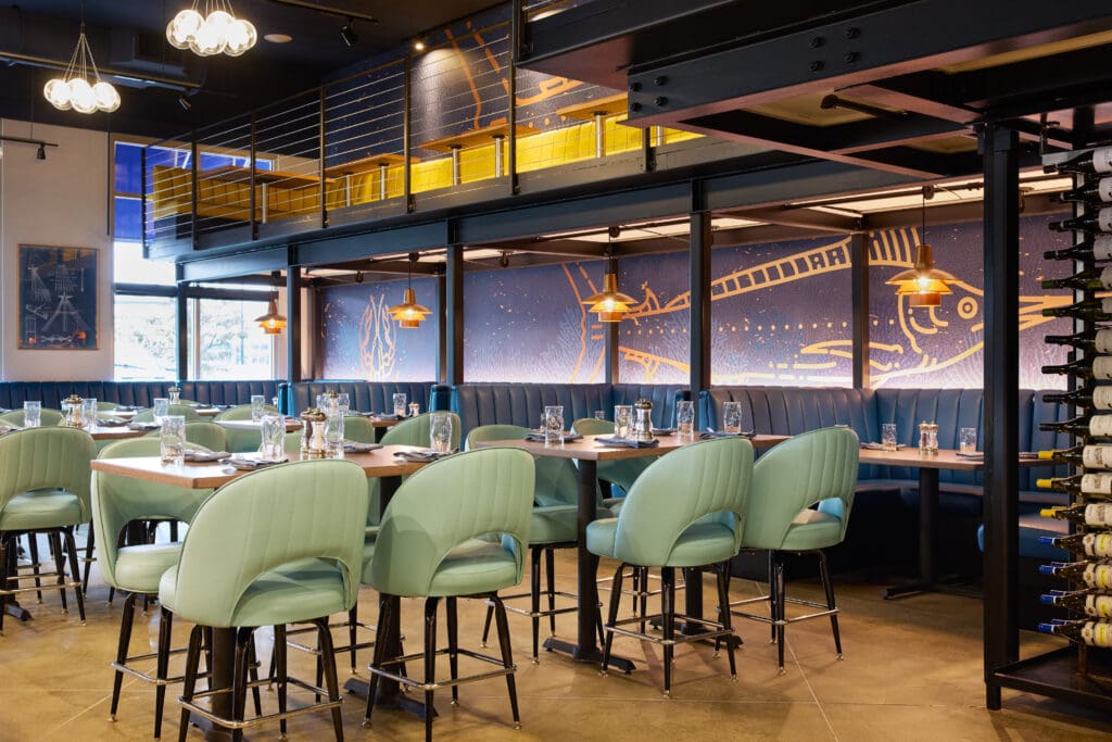 A modern restaurant interior with mint green chairs, wooden tables set with glasses and cutlery, blue cushioned booths, wall art, warm lighting, and a mezzanine level with a yellow railing.