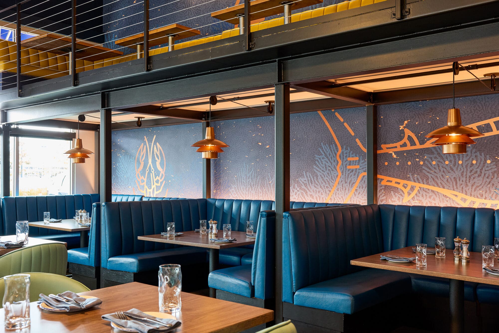 A modern restaurant interior with blue upholstered booths, wooden tables set with glasses and napkins, and copper pendant lights. The wall features ocean-themed artwork with sea creatures in gold outlines.