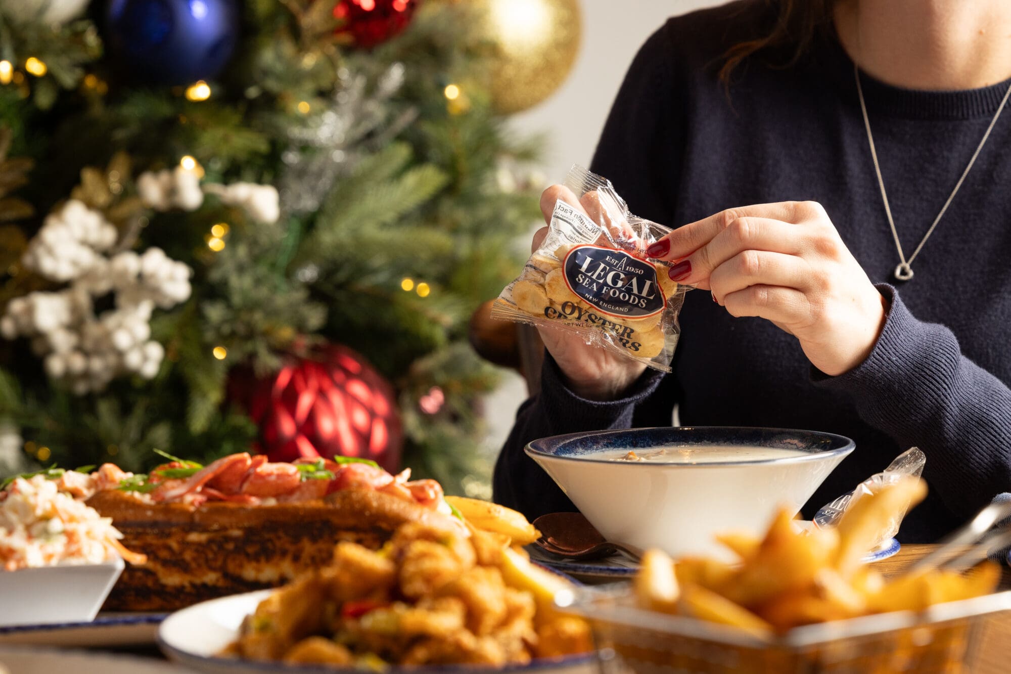 A woman sits at a festive table with holiday decorations, holding a packet of oyster crackers over a bowl of soup. Plates of seafood, fries, and other food are on the table. A Christmas tree is in the background.