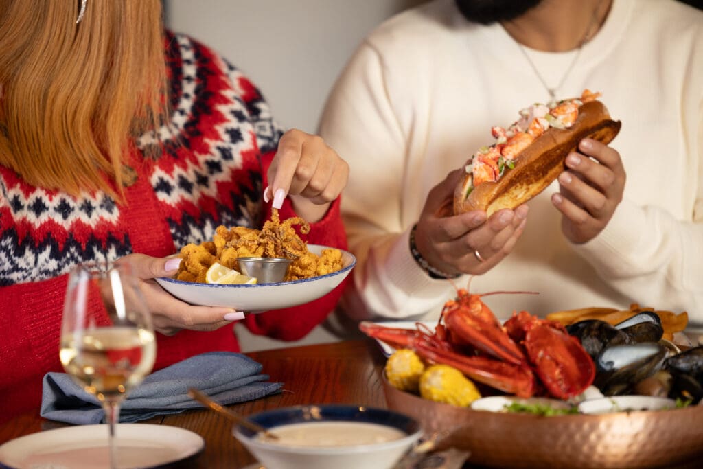 Two people sitting at a table share seafood; one holds a plate of fried calamari, the other holds a lobster roll. A seafood platter, wine glass, and soup are also visible on the wooden table.