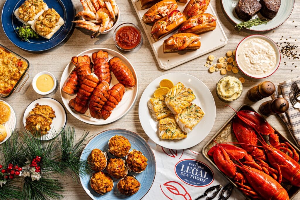 A festive seafood spread featuring lobster tails, shrimp, crab cakes, chowder, stuffed clams, grilled fish, lobster rolls, whole lobsters, sauces, and holiday decor on a wooden table.