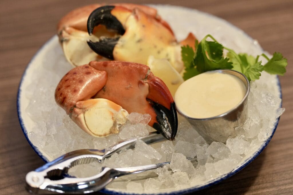 A plate of stone crab claws on crushed ice with a metal cup of creamy dipping sauce, garnished with fresh parsley, and a crab cracker tool on the side.