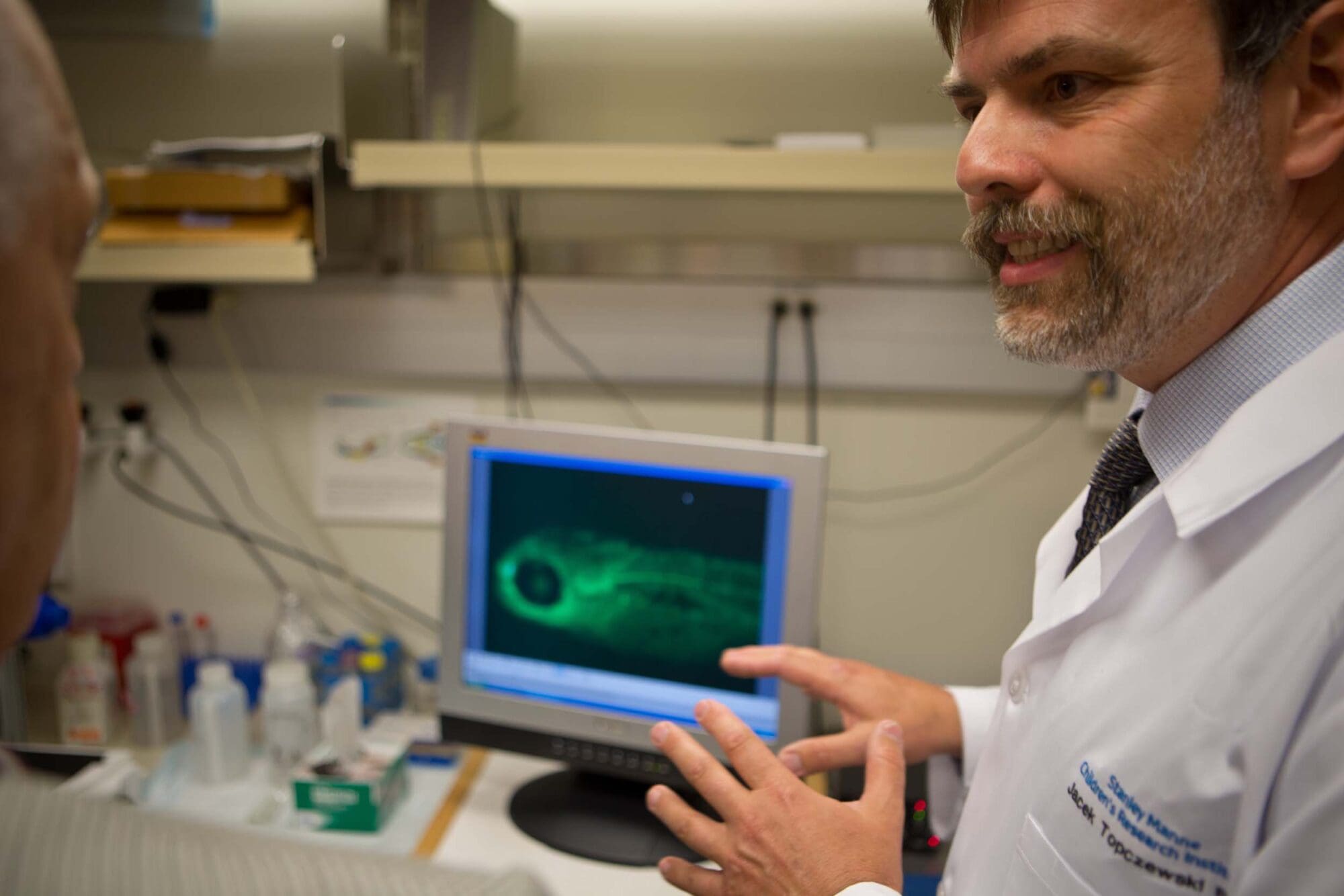 A scientist in a white lab coat gestures while explaining a green fluorescent image of a specimen displayed on a computer monitor in a laboratory setting.