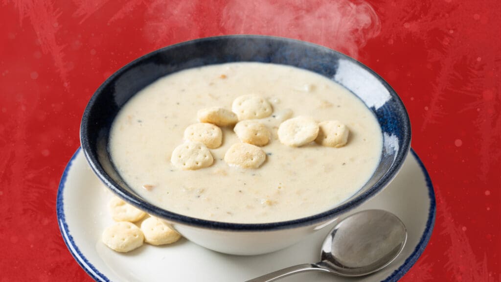 A steaming bowl of creamy soup with oyster crackers on top, served on a white plate with a spoon, against a red background.