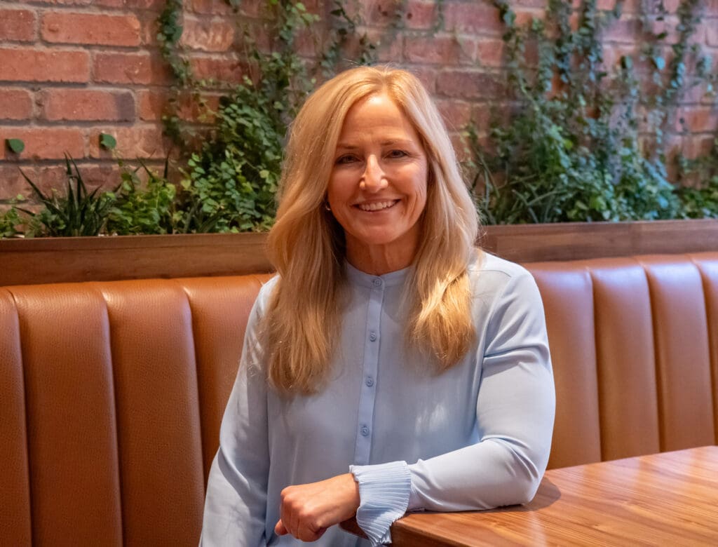 A woman with long blonde hair wearing a light blue blouse sits at a wooden table in a restaurant with brown leather seating and a brick wall with greenery in the background, smiling at the camera.