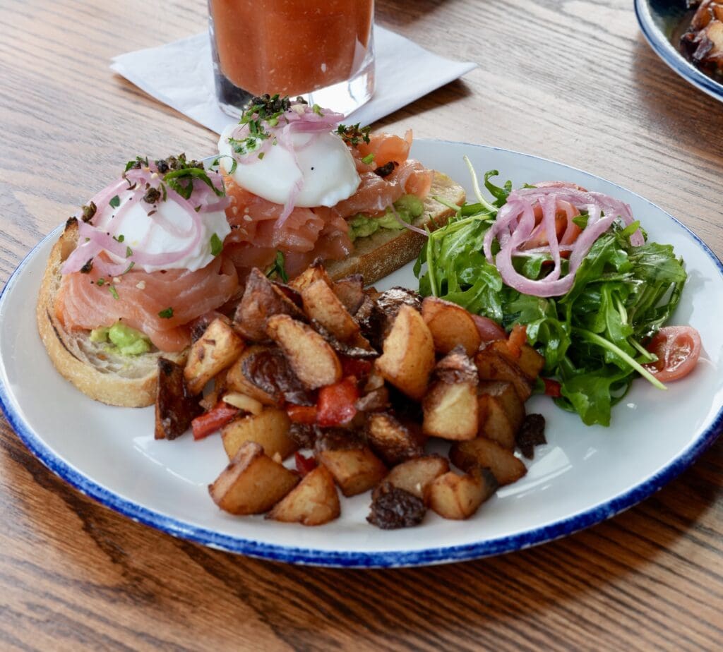 A plate with avocado toast topped with smoked salmon, poached eggs, and pickled onions, served with roasted potatoes and a side salad of arugula and pickled onions. A drink is visible in the background.