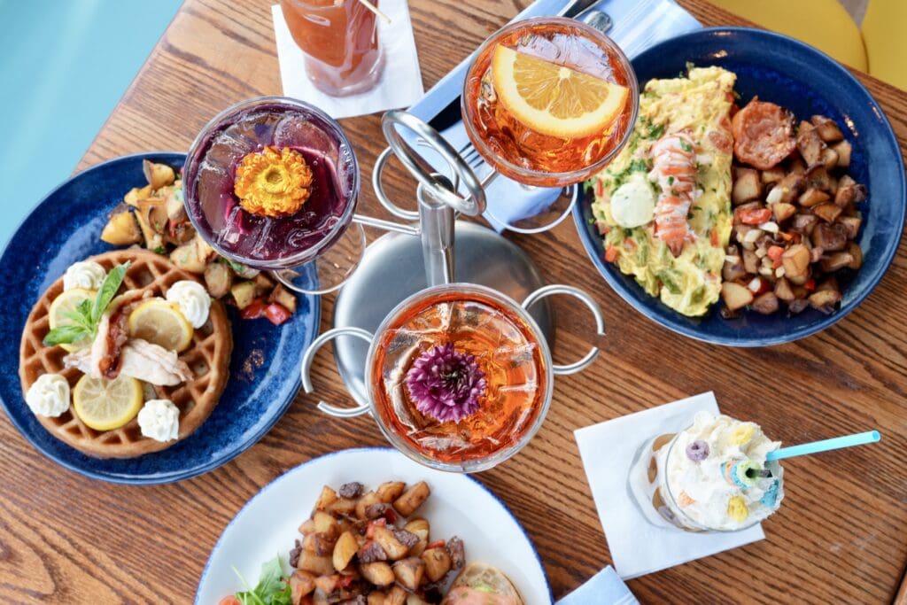 A top-down view of a brunch table with colorful cocktails, a waffle with lemon slices and whipped cream, an omelette with potatoes, a plate of home fries, and a milkshake topped with whipped cream and a cherry.