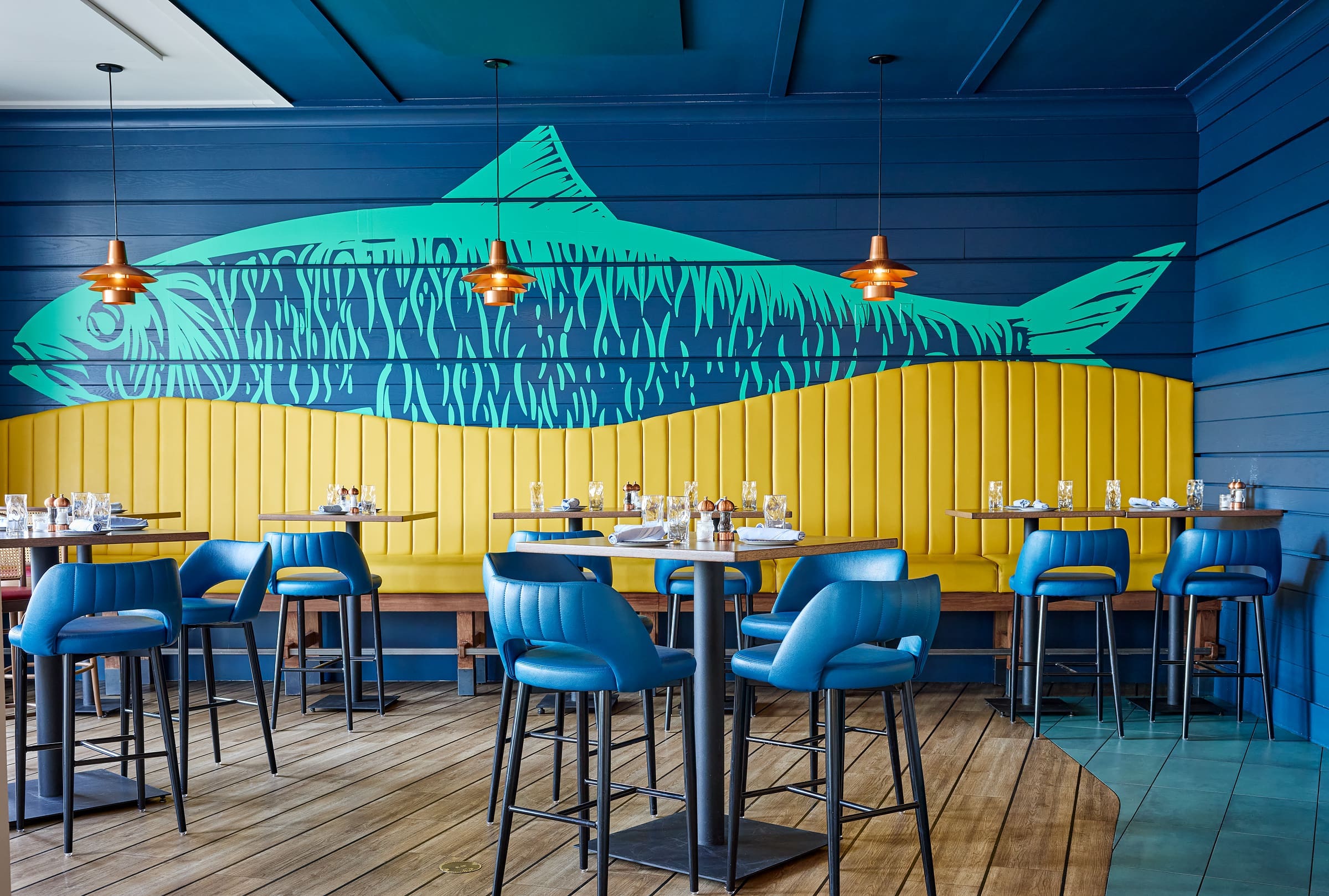 A modern restaurant interior with blue and yellow decor, blue chairs, wooden tables, hanging copper lights, and a large fish mural on the wall behind a yellow bench.