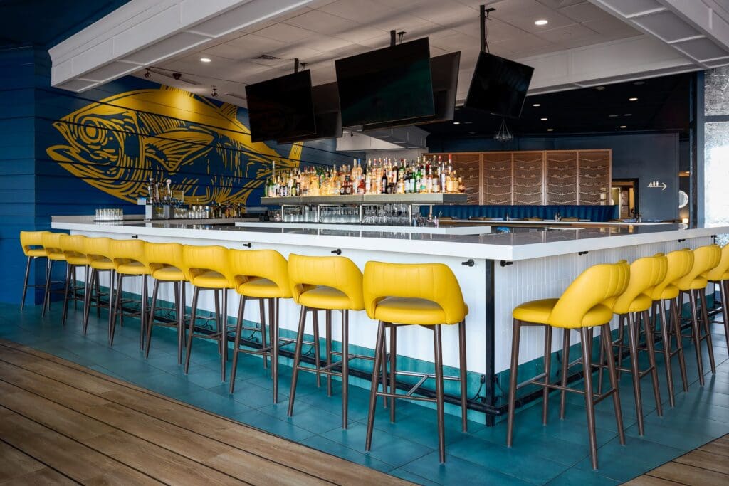 A modern bar with a white countertop and high-backed yellow barstools arranged around it. The background features a large fish mural and shelves stocked with bottles. The bar has blue accents and multiple overhead TVs.