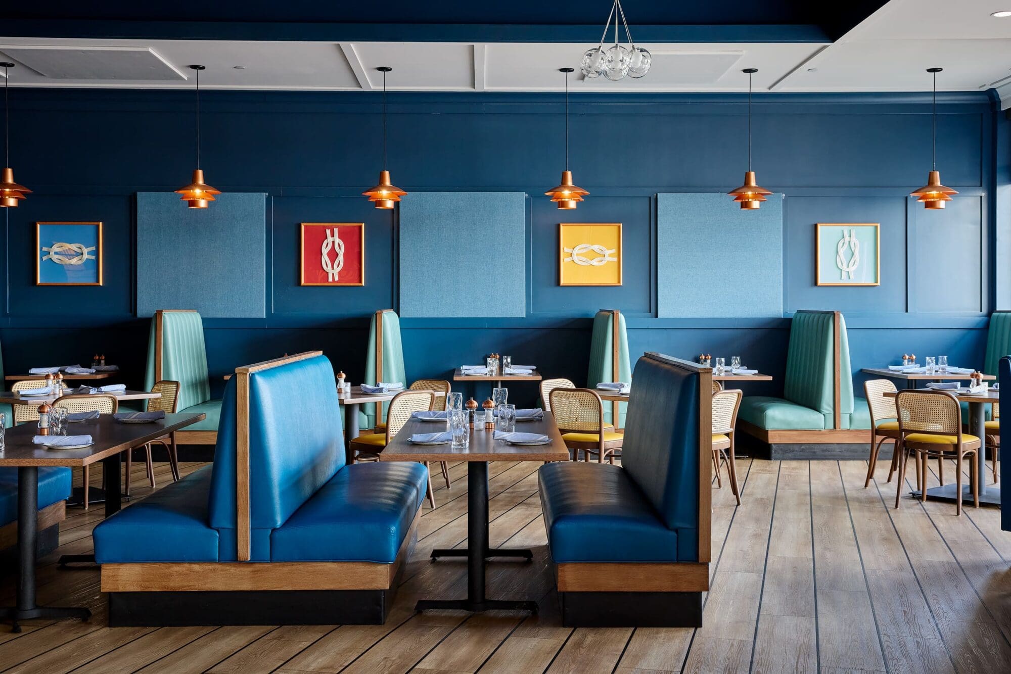 A modern restaurant interior with teal and blue booths, cane-backed chairs, wooden floors, and blue walls decorated with colorful abstract art and copper pendant lights hanging from the ceiling.