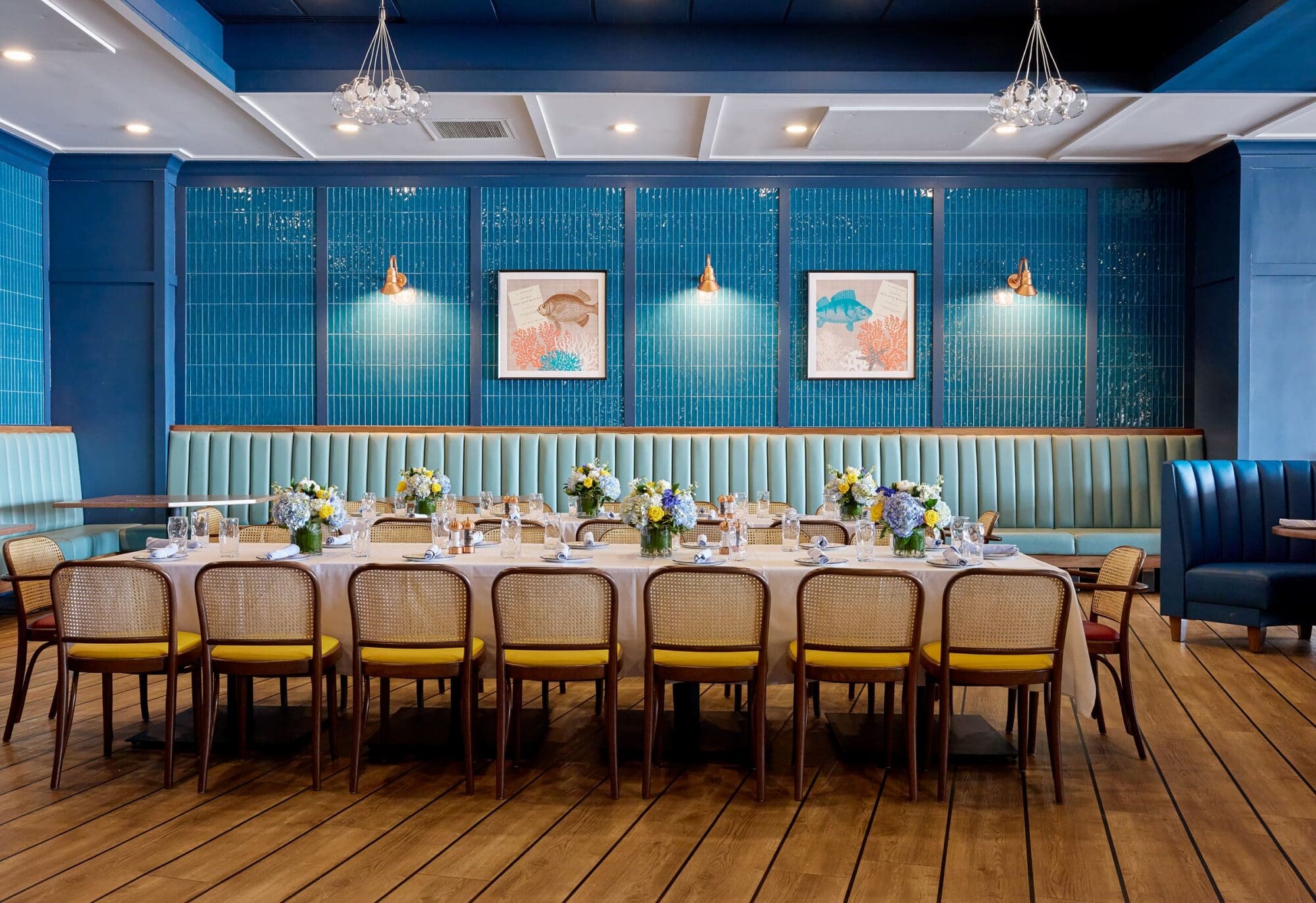 A long dining table with floral centerpieces is set for a meal in a stylish restaurant featuring teal walls, framed art, cushioned banquettes, and wooden chairs on a wood floor. Chandeliers hang from the ceiling above.
