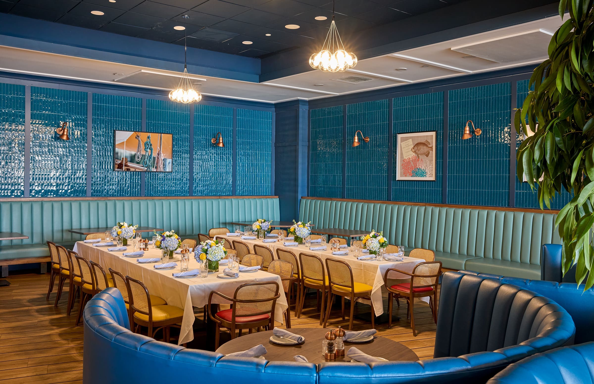 A stylish restaurant interior with teal-blue walls and booths, wooden tables set with white tablecloths, flowers, and place settings. Bright lighting and framed art contribute to the inviting, modern atmosphere.