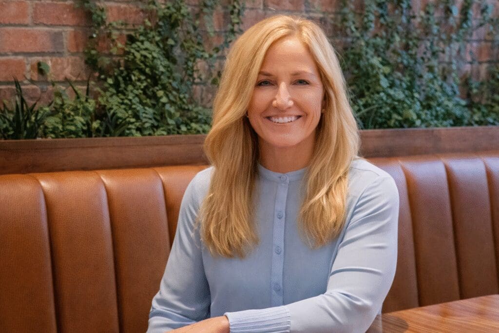 A woman with long blonde hair, wearing a light blue blouse, sits and smiles at a wooden table in a restaurant with brown leather seating and green plants on a brick wall in the background.
