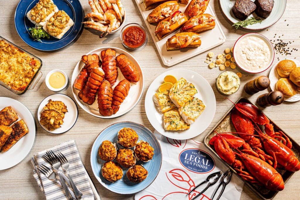 A table filled with assorted seafood dishes, including lobster tails, shrimp, crab cakes, lobster rolls, baked fish, and sides. Legal Sea Foods packaging and utensils are also visible on the table.