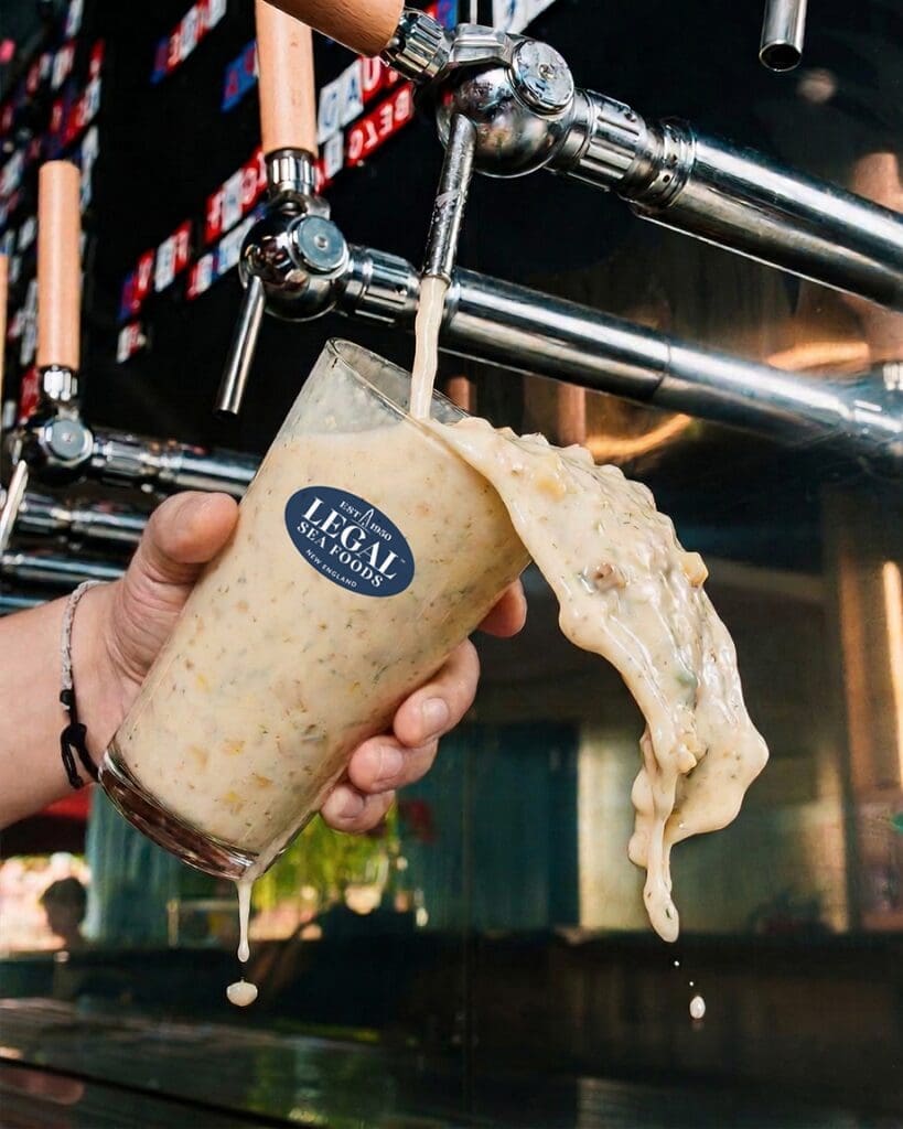 A hand holds a glass of thick clam chowder being poured from a tap, overflowing and spilling down the side. The glass has a 