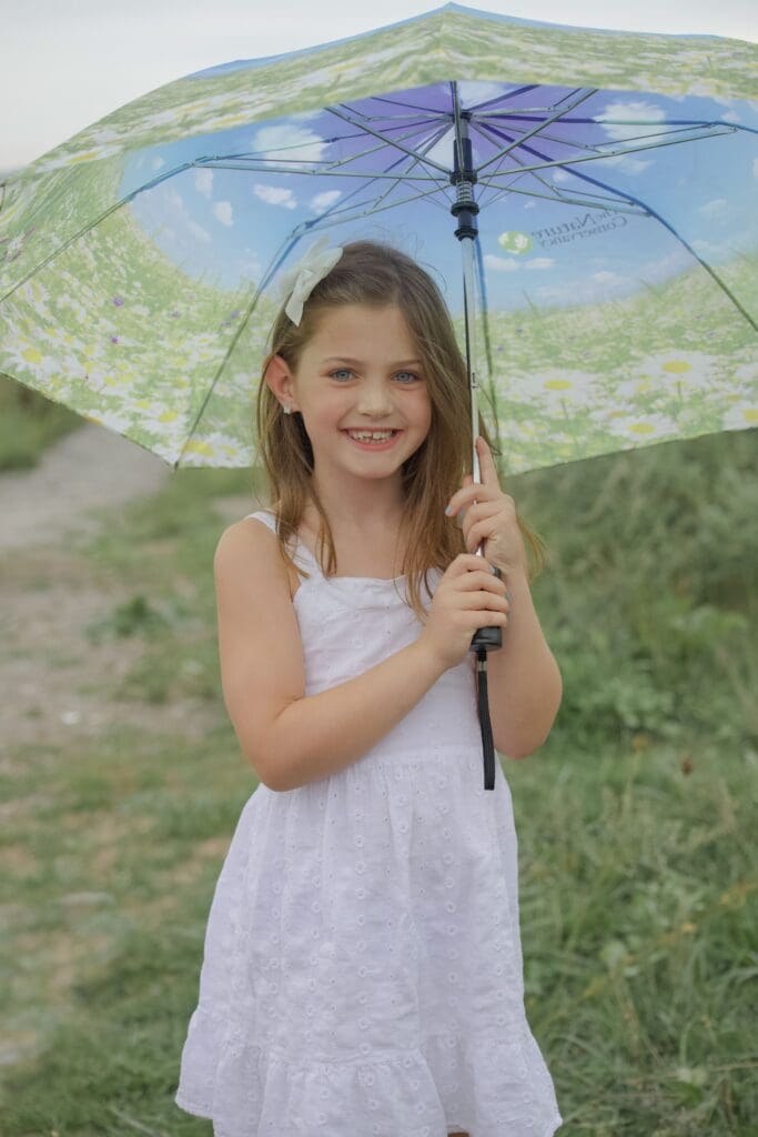 A young girl in a white dress stands outside on a grassy path, smiling and holding a colorful umbrella decorated with a floral pattern and blue sky design.