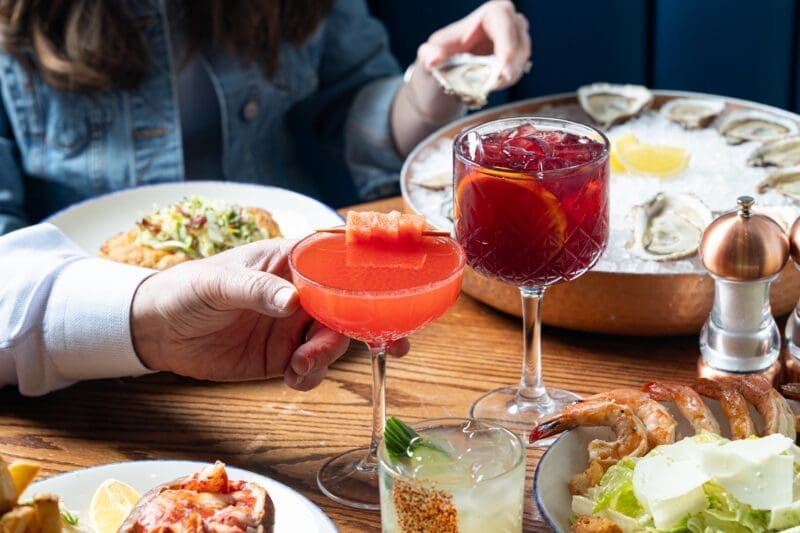 A table with cocktails, oysters on ice, shrimp, and other seafood dishes. Two people are reaching for food and drinks; one holds a pink cocktail, the other holds an oyster.