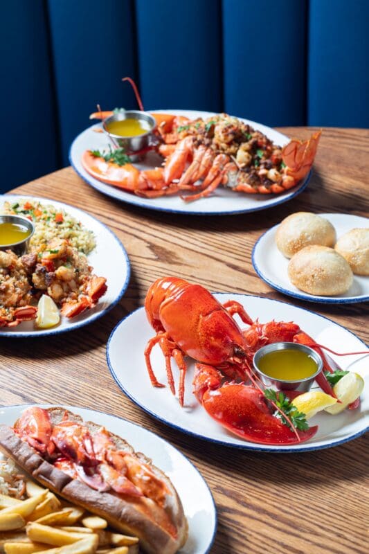 Plates of seafood including whole lobster, lobster roll with fries, shrimp, rice, and bread rolls are arranged on a wooden table with a blue booth in the background. Each dish is served with a side of melted butter.
