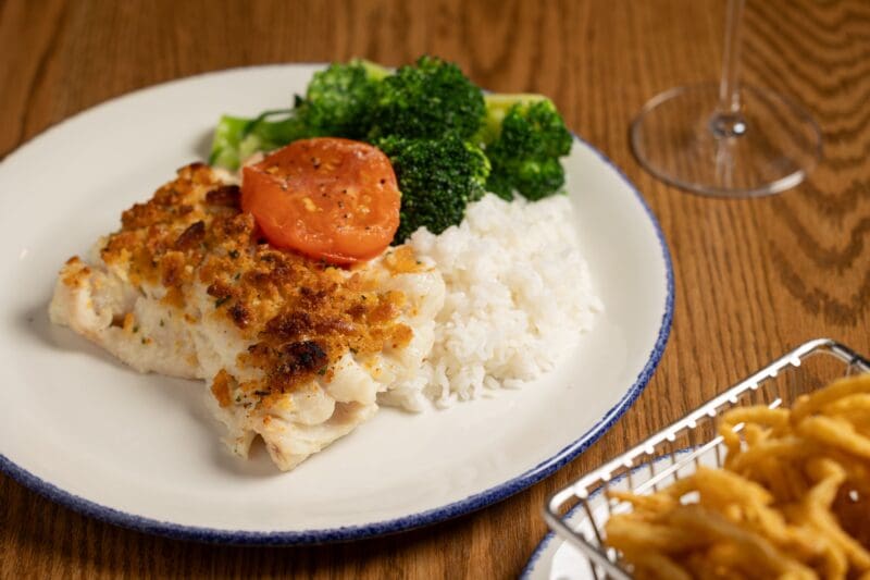 A plate with baked fish topped with a slice of tomato, served with white rice and steamed broccoli. A glass and a basket of fried onion rings are partially visible in the background.