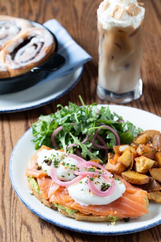 A plate with smoked salmon and poached eggs on toast, topped with pickled onions and chives, served with roasted potatoes and a green salad. In the background, a cinnamon roll and a glass of iced coffee with whipped cream.