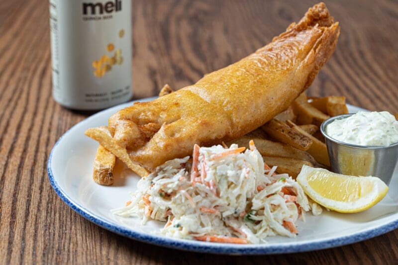 A plate of fish and chips with coleslaw, a lemon wedge, and tartar sauce on the side. A canned drink is in the background, all set on a wooden table.