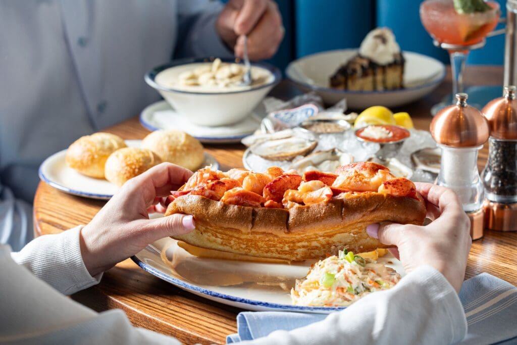 A person holds a lobster roll on a plate with coleslaw, with bowls of soup, bread rolls, oysters, dessert, and a cocktail on the table in a bright, casual dining setting.