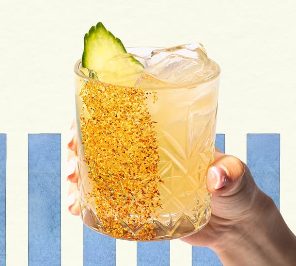 A hand holds a cocktail in a faceted glass with ice, a cucumber slice garnish, and a rim coated in chili powder. Blue vertical stripes and a light background are visible behind the drink.