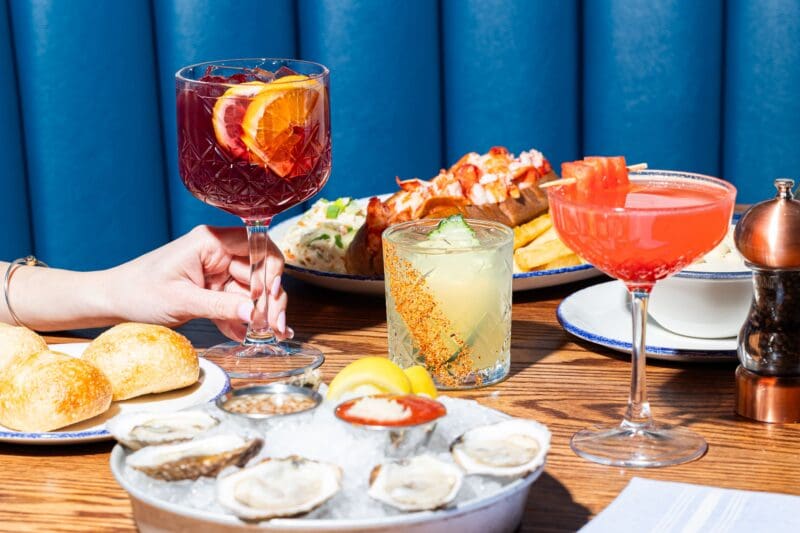 A table set with oysters on ice, bread rolls, and various colorful cocktails. A hand holds a glass of red sangria, with lobster rolls and other drinks in the background, against a blue cushioned booth.