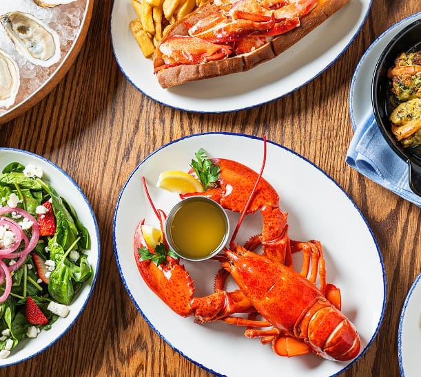 A table set with a whole cooked lobster and melted butter, a lobster roll with fries, a fresh spinach salad with strawberries and feta, oysters on the half shell, and a seafood dish in a blue skillet.