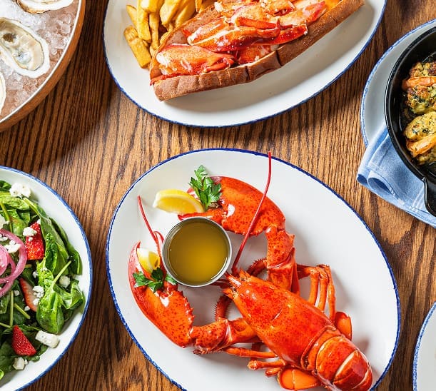 Overhead view of a table with a whole cooked lobster with lemon wedges and butter, a lobster roll with fries, a fresh green salad, a dish of oysters, and a seafood stew on a wooden surface.