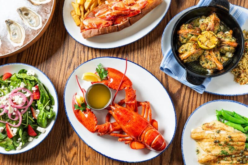A table set with a variety of seafood dishes, including a whole lobster with butter, grilled shrimp, oysters, a lobster roll with fries, a plate of fish with green beans, and a salad with spinach, strawberries, feta, and onions.