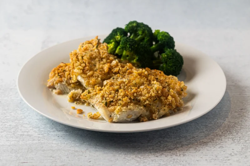 A white plate with two pieces of baked fish topped with a golden, crumbly crust, served alongside a portion of steamed broccoli.