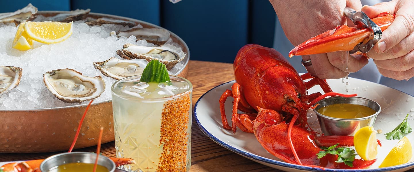 A person cracks open a cooked lobster claw over a plate with melted butter, next to a cocktail and a platter of oysters on ice garnished with lemon wedges.