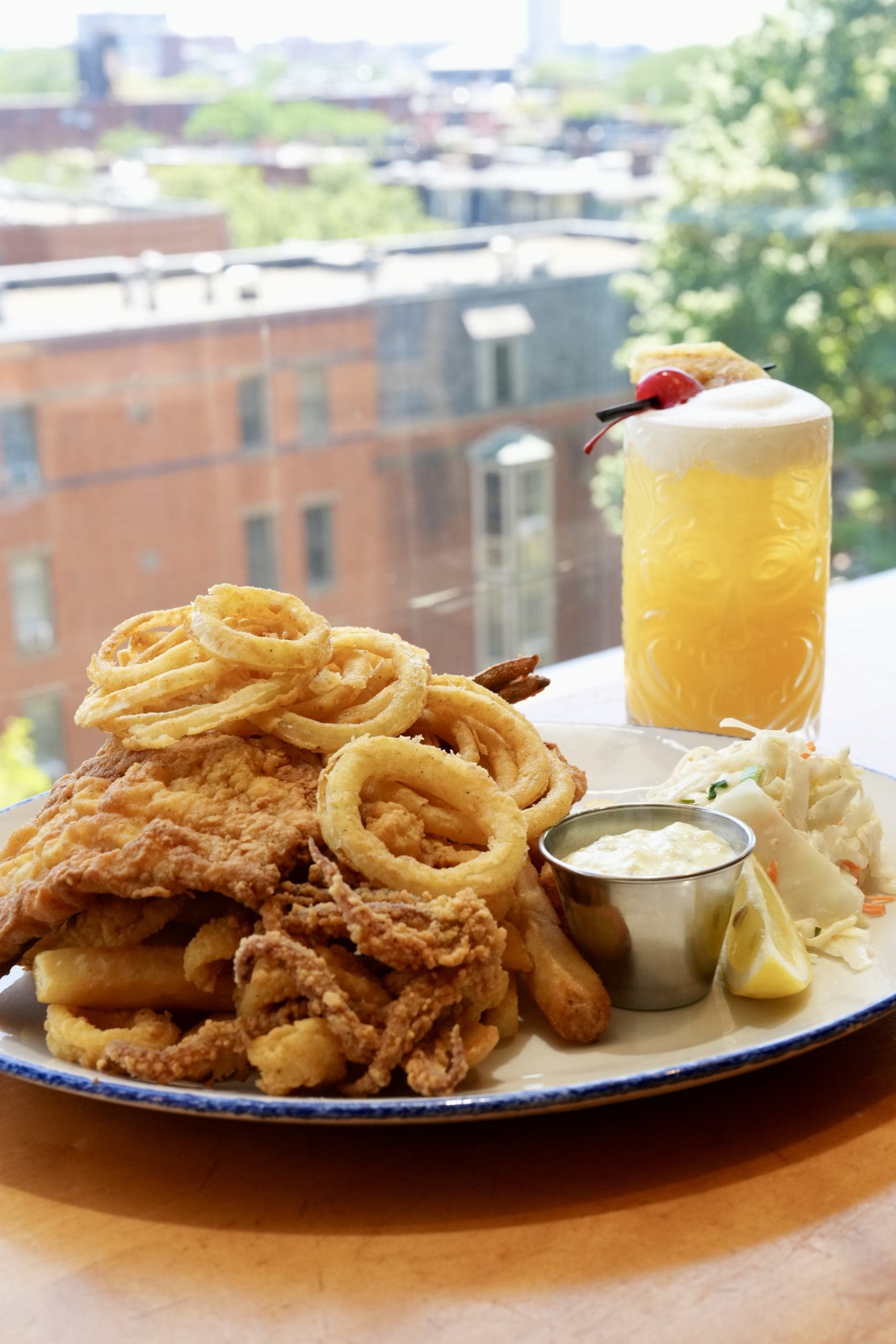 A plate of fried seafood with onion rings, fries, coleslaw, tartar sauce, and a lemon wedge. A yellow drink with foam and a cherry is in the background, with city buildings visible through a window.